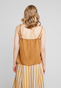 Camisola de satén marrón con tirantes finos, que presenta una parte posterior fruncida. Combinada con una falda a rayas en tonos beige y amarillo.