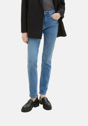 Personne portant un jean slim bleu clair, des mocassins noirs, un blazer noir et un haut gris avec une main dans la poche du jean sur fond blanc.