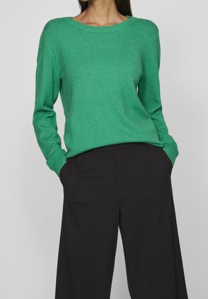 Femme portant un pull en maille vert à manches longues, rentré dans un pantalon noir ample taille haute, les mains dans les poches.