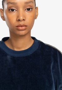 Felpa in velluto blu navy con colletto a costine e dettagli in rilievo sulle spalle. Ingrandimento della superficie del tessuto.