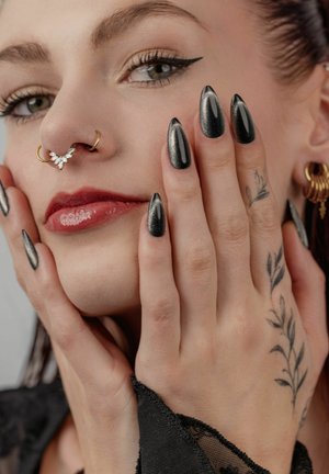 Close-up of a woman with black stiletto nails, floral hand tattoo, gold nose and ear rings, winged eyeliner, and red lipstick touching her face.