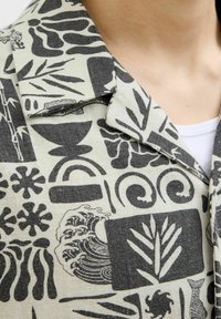 Camisa de manga corta de tela beige con patrones negros, incluyendo olas, plantas y formas abstractas; presenta un cuello estructurado y texturas.
