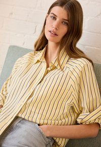 Camicetta button-up a righe gialle con maniche corte, tasca frontale, vestibilità rilassata, abbinata a shorts di jeans chiari, su uno sfondo texturizzato.