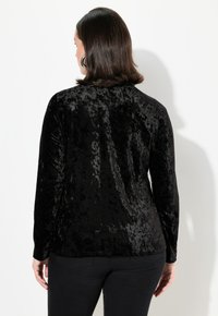 Schwarzes Samtoberteil mit langen Ärmeln und strukturierter Oberfläche, Rundhalsausschnitt und taillierter Passform, kombiniert mit dunklen Hosen.