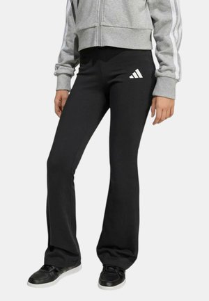 Donna che indossa pantaloni neri a zampa Adidas, giacca grigia con zip e strisce bianche sulle maniche, e sneakers nere con suola bianca.