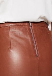 Brauner Lederminirock mit glatter Textur, seitlichem Reißverschluss und sichtbaren Nahtdetails entlang der Naht.