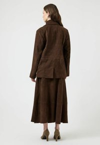 Ruskea mokkainen bleiseri korkealla kauluksella, yhdistettynä vastaavaan polvipituiseen hameeseen. Molemmissa vaatteissa on pehmeä tekstuuri ja vartalonmyötäinen leikkaus.