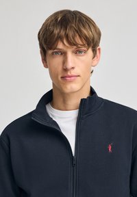 Giacca in pile con zip di colore blu navy e colletto rialzato, con un logo ricamato rosso sul petto e una T-shirt bianca a strati sotto.