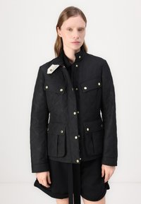Jachetă neagră matlasată cu accesorii aurii, două buzunare frontale și guler în picioare. Prezintă un accent alb contrastant pe umăr.
