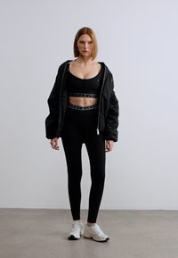 Μαύρο μπουφάν μπόμπερ, αθλητικό σουτιέν και κολάν. Διαθέτει ελαστική ζώνη μέσης και λογότυπο. Το μοντέλο φοράει ανοιχτόχρωμα αθλητικά παπούτσια.