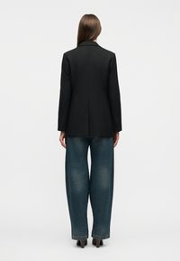 Γυναίκα με μακριά καστανά μαλλιά που φοράει μαύρο σακάκι, μπλε ξεβαμμένο τζιν φαρδιάς γραμμής και καφέ παπούτσια με τακούνι, στέκεται γυρισμένη προς τα πίσω σε απλό φόντο.