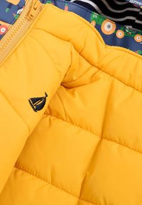 Intérieur d'une veste à douille jaune avec un motif matelassé. Présente une doublure à motifs bleus et un petit logo brodé d'un voilier.