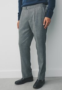 Grau gemusterte Hose mit taillierter Passform, flachem Front, Knopfverschluss und Seitenverstellern, kombiniert mit schwarzen Slippern.