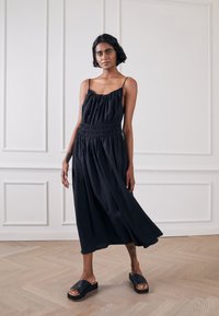 Vestido preto sem mangas com corpete franzido, cintura canelada e saia fluida. Combinado com sandálias de plataforma pretas sobre um chão de madeira.