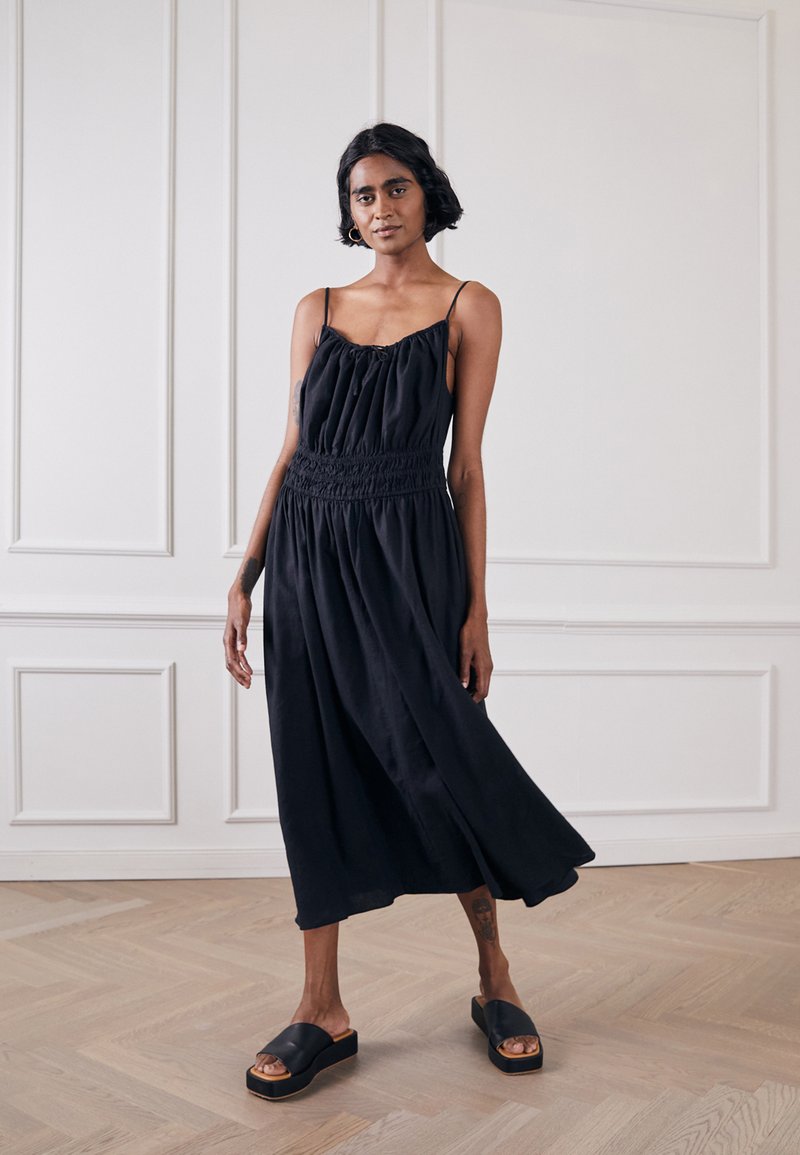 Vestido preto sem mangas com corpete franzido, cintura canelada e saia fluida. Combinado com sandálias de plataforma pretas sobre um chão de madeira.