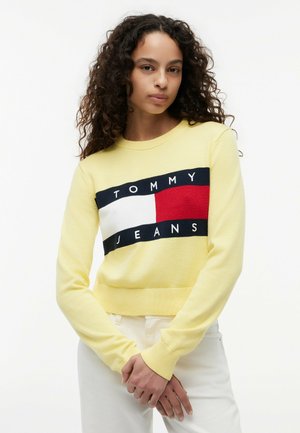 Kvinde med langt krøllet hår iført en gul sweater med et rødt, hvidt og sort Tommy Jeans-logo og hvide bukser, står med hænderne foldet.