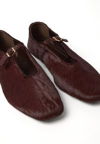 Un paio di scarpe slip-on in pelliccia marrone scuro con fibbie dorate e punte arrotondate su uno sfondo bianco.