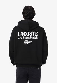 Persona con una sudadera con capucha negra que tiene en la parte posterior el texto blanco "LACOSTE Jeu Set et Match" y un logo de cocodrilo.