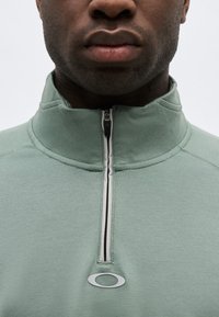 Κοντινό πλάνο ενός ατόμου που φοράει ανοιχτό πράσινο τοπ με φερμουάρ και όρθιο γιακά, με μικρό λογότυπο Oakley κάτω από το φερμουάρ.