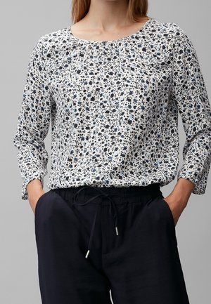 Femme portant un chemisier blanc à manches longues à petits motifs floraux noirs et bleus, rentré dans un pantalon noir à cordon de serrage, mains dans les poches.