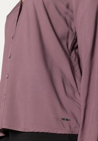 Acercamiento de una camisa de botón en color malva llevada sobre pantalones negros, mostrando la costura lateral y una pequeña etiqueta de metal de la marca cerca del dobladillo.