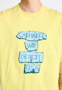Kollane puuvillane t-särk, millel on graafiline disain sinise, kulunud tekstiga, mis ütleb "Carhartt WIP Detroit USA" kihiliselt plokk-kujudes.