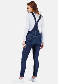 Denim tuinbroeken met verstelbare banden, donkerblauwe was, aansluitende vorm, achterzakken en een logo-patch op de taille. Gecombineerd met witte schoenen.