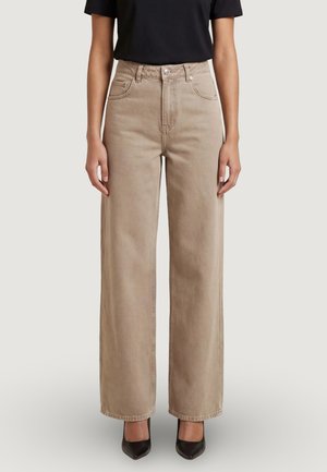 Donna che indossa pantaloni beige a vita alta e gamba larga, tacchi neri con punta a punta e una maglietta nera a maniche corte, in piedi davanti a uno sfondo semplice.