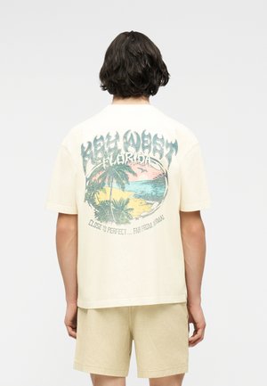 Person mit dunklem lockigem Haar steht mit dem Rücken zur Kamera, trägt ein cremefarbenes Hemd mit einem bunten Key West Florida Strandmotiv und beigen Shorts.