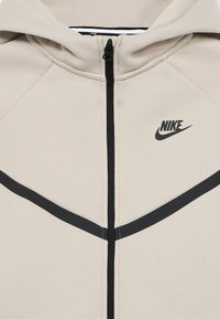 Ελαφρώς μπεζ φούτερ με κουκούλα, μαύρες λεπτομέρειες και φερμουάρ μπροστά. Διαθέτει λογότυπο Nike, ραβδωτή υφή και κουκούλα. Σχεδιασμός με οριζόντιες μαύρες ρίγες.