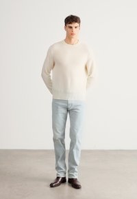 Gaiši bēšs adīts džemperis ar vaļēju piegriezumu un ribām piedurknēs, kombinēts ar gaiši zilām džinsu biksēm un tumši brūnām ādas kurpēm. Vienkāršs, minimālistisks stils.