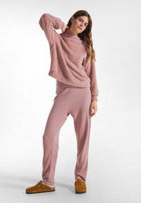 Pullover rosa e peloso con colletto alto e maniche ampie abbinato a pantaloni larghi coordinati; il modello indossa zoccoli marroni. Texture morbida nel complesso.