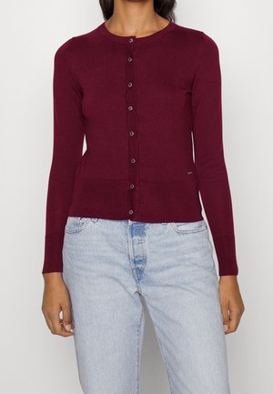 Frau trägt eine taillierte, langärmelige, geknöpfte burgunderrote Strickjacke kombiniert mit hellblauen, hoch taillierten Jeans vor neutralem Hintergrund.