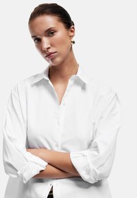 Camisa blanca de botón con un cuello clásico, mangas enrolladas y una textura suave. Presenta una sola fila de botones en la parte delantera.
