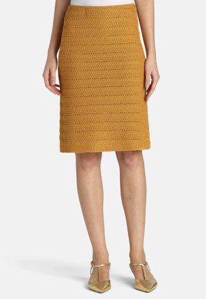 Knedlang karrygul tekstureret nederdel båret med guldspidsede hæle og hvid top, vist fra talje til fødder.