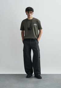 T-shirt vert olive avec logo, pantalon cargo noir avec poches latérales et baskets noires. Les accessoires comprennent des lunettes de soleil rondes et un porte-né.