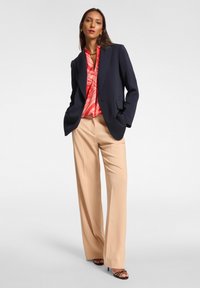 Blazer navy sopra una blusa rossa a fantasia, abbinato a pantaloni beige a gamba larga. Presenta una trama liscia e dettagli hardware minimi.