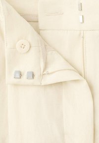 Gros plan d’un pantalon beige clair montrant un bouton, une fermeture éclair et une fermeture à crochet et barre métallique sur la ceinture en tissu et la braguette.