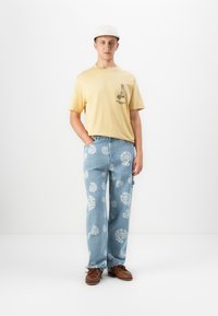Ung mand iført en beige kasket, gult grafisk T-shirt, lyseblå jeans med hvide spiral mønstre og brune lædersko, stående op ad en hvid væg.