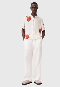 Hombre de pie con las manos en los bolsillos, vistiendo pantalones anchos blancos, camiseta blanca de manga corta con estampados de rosas rojas y zapatos beige.