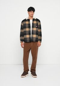 Plaid jacket in black, beige, and brown hues with a cream collar, button front, and side pockets. Paired with brown corduroy pants and sneakers.