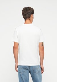 White cotton t-shirt with a crew neckline, short sleeves, and a relaxed fit. Visible tattoo on the left arm. Worn with blue denim jeans.