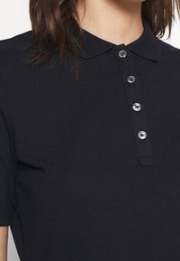 Femme portant un polo noir à manches courtes avec un col et quatre boutons en argent visibles sur le devant.