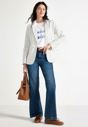 Donna che indossa un blazer bianco a righe sottili, una t-shirt bianca con grafica, jeans blu a gamba larga, scarpe di pelle marrone e porta una borsa di pelle marrone.