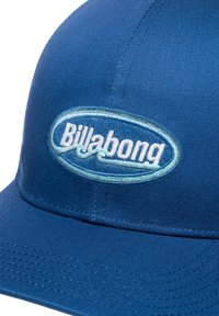Cappellino blu realizzato in tessuto testurizzato; presenta un logo bianco ricamato "Billabong" all'interno di un ovale aqua. Visiera curva con parte inferiore rigata.