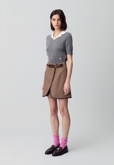 Una donna sta in piedi con sicurezza, indossando un maglione grigio a maniche corte, una gonna a quadri marrone con una cintura e calzini rosa con mocassini.