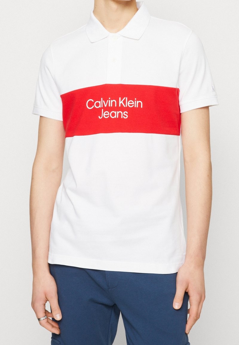 Koszulka polo z krótkim rękawem w kolorze białym z czerwoną poziomą paską, z białym logo "Calvin Klein Jeans". Materiał bawełniany, klasyczny krój kołnierzyka.