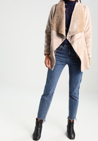 Béžová bunda z umělé semiše s volně padajícím límcem, doplněná modrými džíny s vysokým pasem a černými kotníkovými botami.