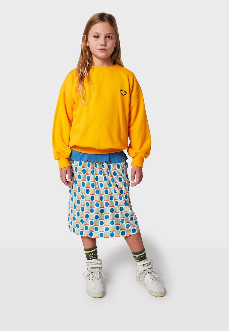Felpa gialla, sottogiacca blu, gonna midi a fantasia con cerchi multicolori, sneakers alte bianche, calzini verdi con testo.