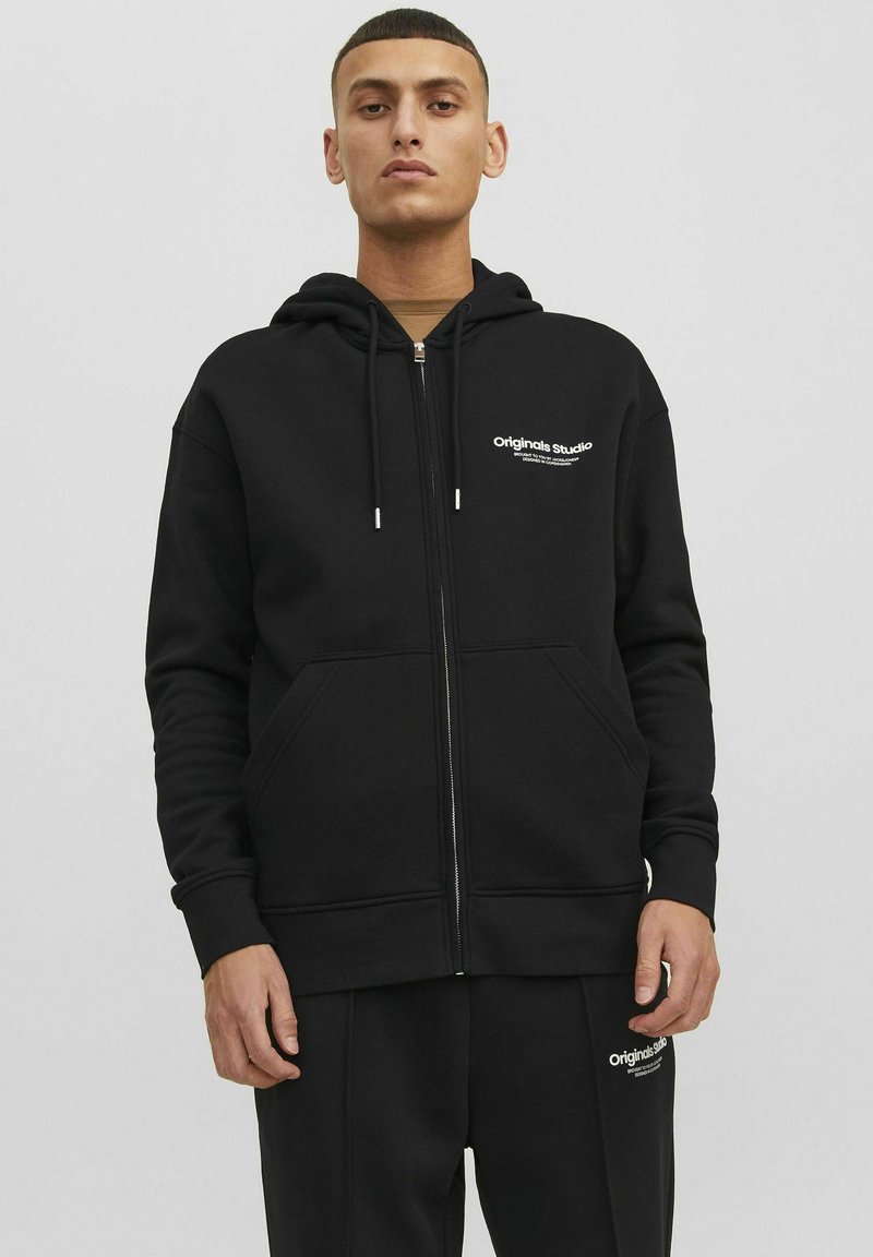 Zwarte zip-up hoodie van zacht materiaal, met een trekkoordcapuchon, zijzakken en een wit logo op de borst.